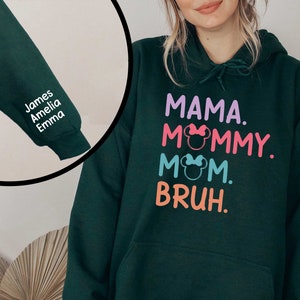 K&ouml;nnte beinhalten: Ein dunkelgr&uuml;ner Hoodie mit einem wei&szlig;en Aufdruck, der "MAMA. MOMMY. MOM. BRUH." mit einem rosa, blauen und orangenen Farbschema sagt. Der Hoodie hat eine Tasche auf der Vorderseite und eine Kapuze mit einem Kordelzug. Der &Auml;rmel hat einen wei&szlig;en Aufdruck mit den Namen "James, Amelia, Emma".