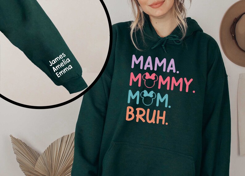 K&ouml;nnte beinhalten: Ein dunkelgr&uuml;ner Hoodie mit einem wei&szlig;en Aufdruck, der "MAMA. MOMMY. MOM. BRUH." mit einem rosa, blauen und orangenen Farbschema sagt. Der Hoodie hat eine Tasche auf der Vorderseite und eine Kapuze mit einem Kordelzug. Der &Auml;rmel hat einen wei&szlig;en Aufdruck mit den Namen "James, Amelia, Emma".
