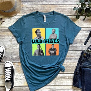 May include: Teal blue t-shirt featuring a graphic of four men in black and white photographs. The photos are in squares with coloured backgrounds. The text "DAD VIBES" is in a retro font.