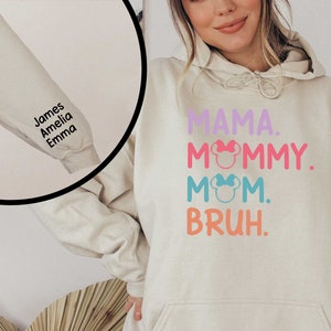 K&ouml;nnte beinhalten: Ein hellbeiger Hoodie mit dem Text "MAMA. MOMMY. MOM. BRUH." in Rosa, Blau und Orange. Der Text ist mit kleinen rosa und blauen Schleifen verziert. Der Hoodie hat einen personalisierten &Auml;rmel mit den Namen "James, Amelia, Emma" in schwarzem Text.