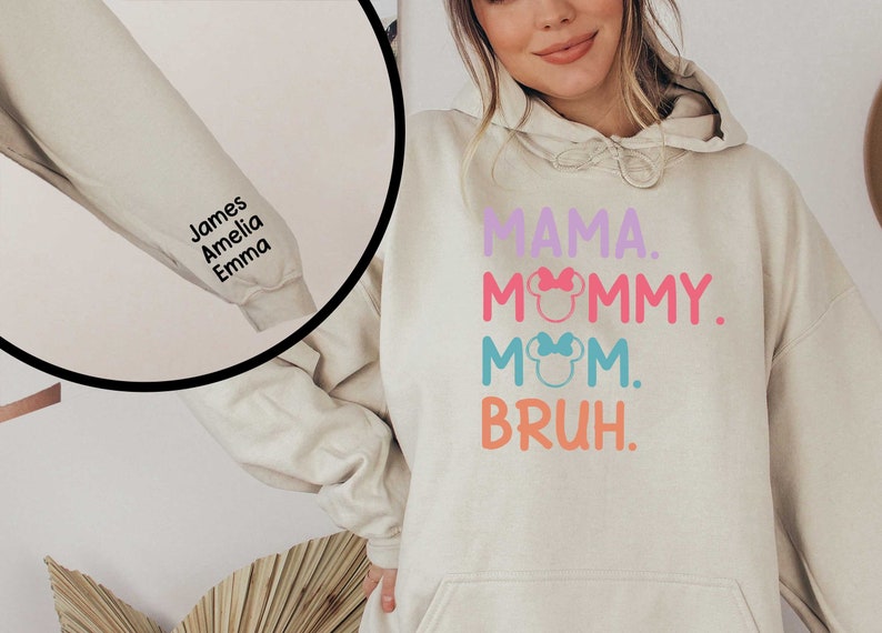 K&ouml;nnte beinhalten: Ein hellbeiger Hoodie mit dem Text "MAMA. MOMMY. MOM. BRUH." in Rosa, Blau und Orange. Der Text ist mit kleinen rosa und blauen Schleifen verziert. Der Hoodie hat einen personalisierten &Auml;rmel mit den Namen "James, Amelia, Emma" in schwarzem Text.