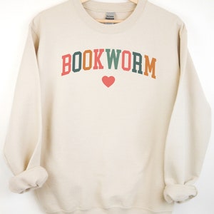 Puede incluir: Una sudadera de color crema con el texto "BOOKWORM" en letras de colores con un coraz&oacute;n rojo debajo.