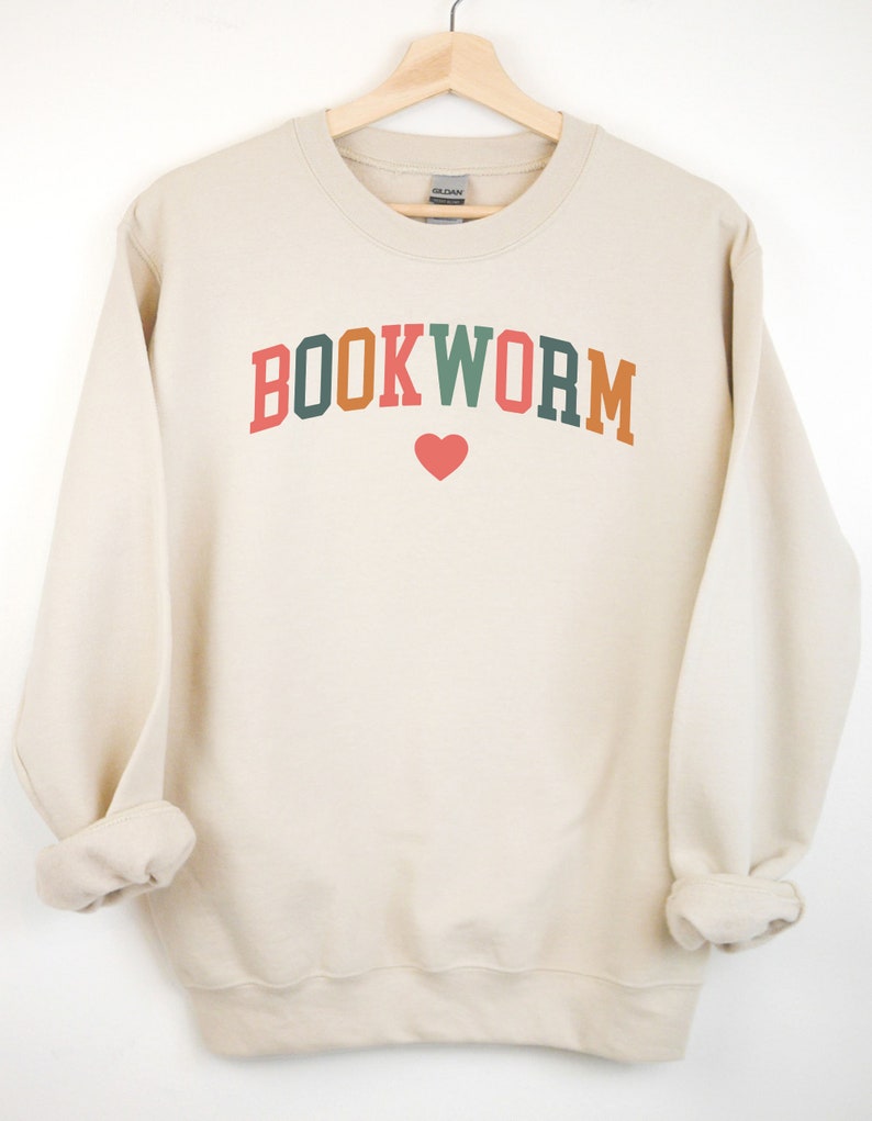 Puede incluir: Una sudadera de color crema con el texto "BOOKWORM" en letras de colores con un coraz&oacute;n rojo debajo.
