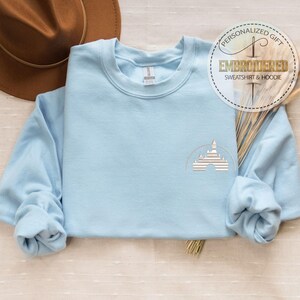 Puede incluir: Sudadera azul claro con un contorno bordado blanco de la silueta del castillo de Cenicienta. La sudadera tiene cuello redondo y mangas largas. El texto "Regalo personalizado Sudadera y sudadera con capucha bordada" est&aacute; en un c&iacute;rculo blanco con un contorno dorado.