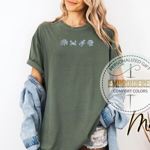 May include: Moss green t-shirt with embroidered sea life designs: seaweed, crab, lobster, and seashell. The shirt has rolled sleeves and a relaxed fit. The text "PERSONALIZED GIFT" and "EMBROIDERED COMFORT COLORS" are printed on a circular label.