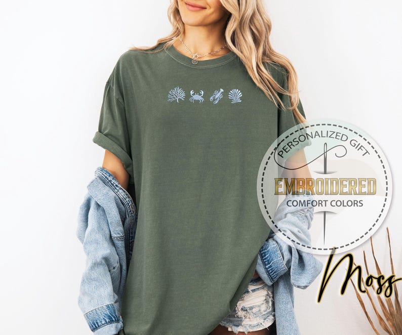 May include: Moss green t-shirt with embroidered sea life designs: seaweed, crab, lobster, and seashell. The shirt has rolled sleeves and a relaxed fit. The text "PERSONALIZED GIFT" and "EMBROIDERED COMFORT COLORS" are printed on a circular label.