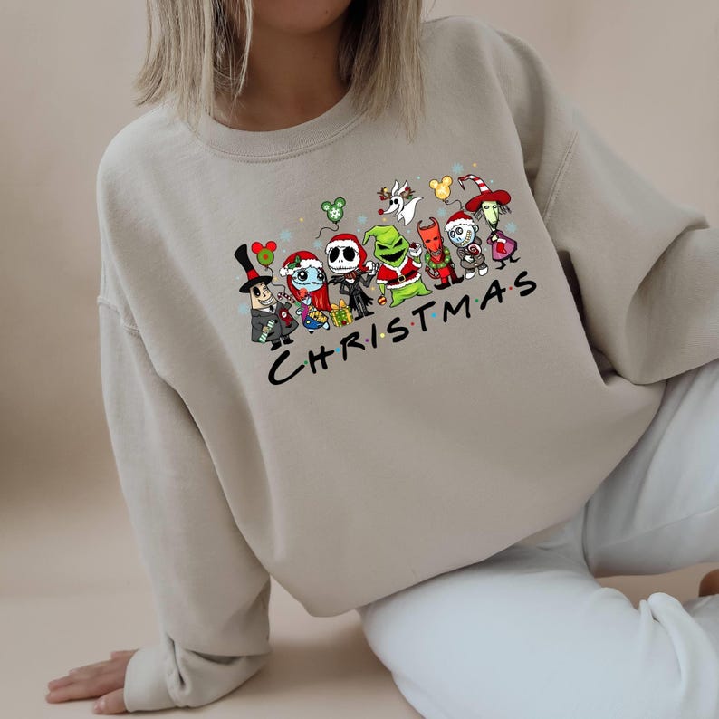 Pu&ograve; includere: Una felpa beige chiaro con una stampa grafica di personaggi dei cartoni animati di film di Natale popolari. I personaggi si tengono per mano e il testo "CHRISTMAS" &egrave; stampato sotto.