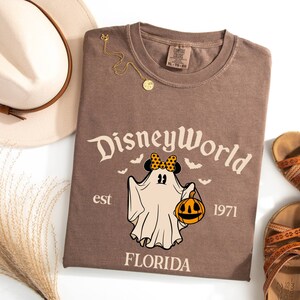 May include: Brown t-shirt featuring a vintage-style graphic of a ghost wearing a Minnie Mouse bow and holding a pumpkin. The text "Disney World est 1971 Florida" is printed on the shirt.