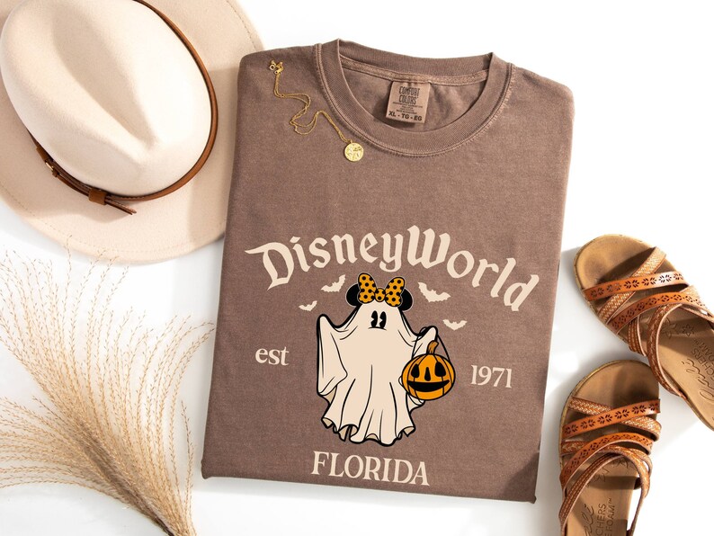 May include: Brown t-shirt featuring a vintage-style graphic of a ghost wearing a Minnie Mouse bow and holding a pumpkin. The text "Disney World est 1971 Florida" is printed on the shirt.