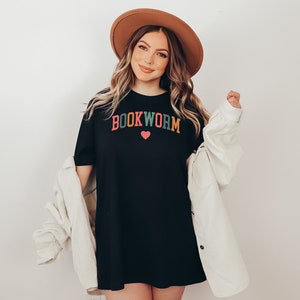 Puede incluir: Una camiseta negra de gran tama&ntilde;o con el texto "BOOKWORM" en letras de colores y un peque&ntilde;o coraz&oacute;n rosa. La modelo lleva un sombrero fedora marr&oacute;n y una chaqueta de pana blanca.