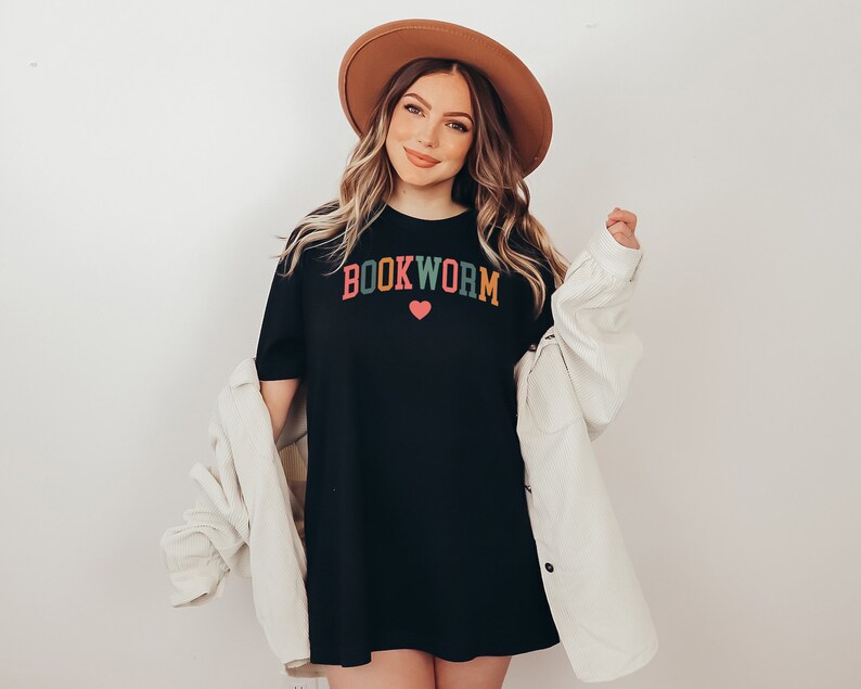 Puede incluir: Una camiseta negra de gran tama&ntilde;o con el texto "BOOKWORM" en letras de colores y un peque&ntilde;o coraz&oacute;n rosa. La modelo lleva un sombrero fedora marr&oacute;n y una chaqueta de pana blanca.