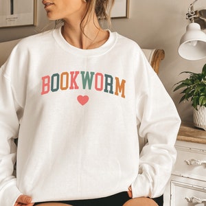Puede incluir: Sudadera blanca con el texto "BOOKWORM" en letras de colores con un coraz&oacute;n rosa en el medio.