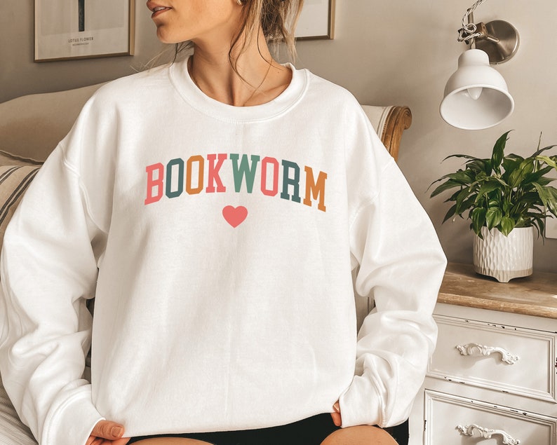 Puede incluir: Sudadera blanca con el texto "BOOKWORM" en letras de colores con un coraz&oacute;n rosa en el medio.