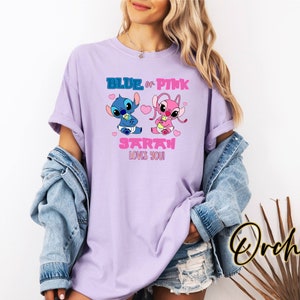 May include: A lilac-colored t-shirt featuring cartoon characters Stitch and Angel, with the text "BLUE or PINK SARAH LOVES YOU!" in blue and pink. The design includes hearts and baby-themed elements. The shirt is part of a clothing collection.