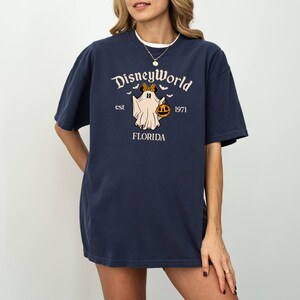 May include: A navy blue t-shirt featuring a white graphic of a ghost wearing a Minnie Mouse bow and holding a pumpkin. The text "Disney World est 1971 Florida" is printed on the shirt.