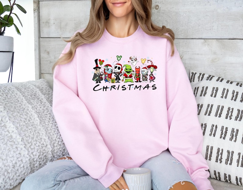 Pu&ograve; includere: Felpa rosa con una grafica di vari personaggi dei cartoni animati di film di Natale popolari. I personaggi sono in piedi insieme e il testo "Christmas" &egrave; stampato sotto di loro.