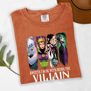 Pu&ograve; includere: T-shirt arancione bruciato con quattro personaggi di cattivi dei cartoni animati. La maglietta ha il testo bianco "Sweetie I'm ok with being the Villain". La maglietta &egrave; piegata su una superficie bianca.