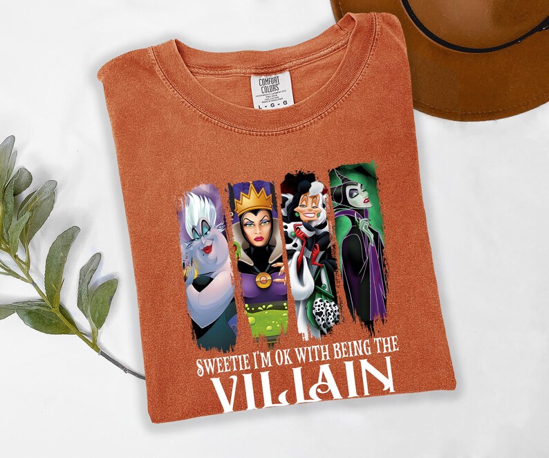 Pu&ograve; includere: T-shirt arancione bruciato con quattro personaggi di cattivi dei cartoni animati. La maglietta ha il testo bianco "Sweetie I'm ok with being the Villain". La maglietta &egrave; piegata su una superficie bianca.