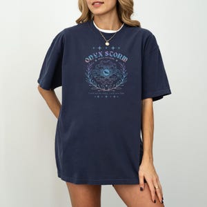 May include: A navy blue oversized t-shirt with a graphic print of a stylized eye with clouds and leaves. The text "ONYX STORM" is above the eye and the text "WE BRAVE THE DARK" is below the eye. The text "I will not fear for today, I will save him." is below the graphic.
