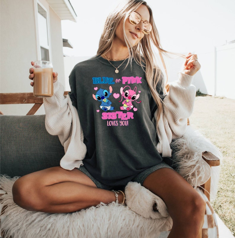 May include: Dark gray unisex t-shirt featuring a graphic of two cartoon characters, one blue and one pink, with the text "BLUE or PINK SISTER LOVES YOU!". The design includes hearts and baby-themed elements.