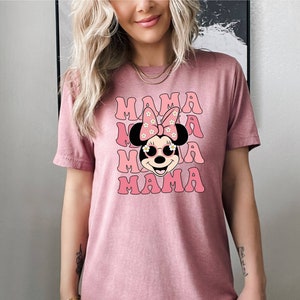 Pu&ograve; includere: Maglietta rosa con un'immagine di un personaggio dei cartoni animati con un fiocco rosa e occhiali da sole. Il testo "MAMA" &egrave; ripetuto quattro volte in un font retr&ograve;.