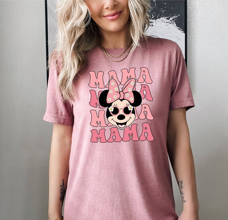 Pu&ograve; includere: Maglietta rosa con un'immagine di un personaggio dei cartoni animati con un fiocco rosa e occhiali da sole. Il testo "MAMA" &egrave; ripetuto quattro volte in un font retr&ograve;.