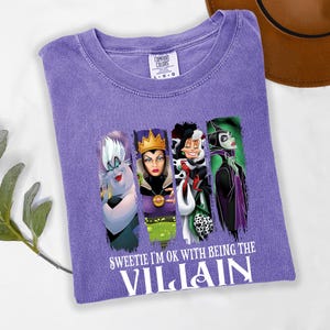 Pu&ograve; includere: Una maglietta viola Comfort Colors con quattro cattivi dei cartoni animati e la scritta "Sweetie I'm ok with being the Villain". La maglietta ha un girocollo e maniche corte. Un cappello marrone &egrave; sullo sfondo.
