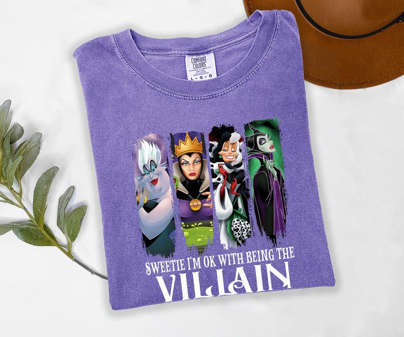 Pu&ograve; includere: Una maglietta viola Comfort Colors con quattro cattivi dei cartoni animati e la scritta "Sweetie I'm ok with being the Villain". La maglietta ha un girocollo e maniche corte. Un cappello marrone &egrave; sullo sfondo.