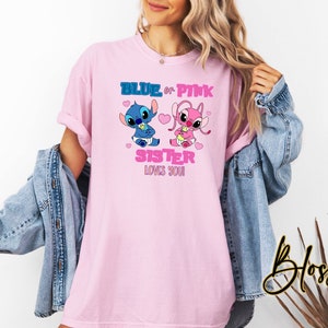 May include: Pink t-shirt featuring two cartoon characters, one blue and one pink, with the text "BLUE or PINK SISTER LOVES YOU!" The design includes hearts and baby-themed elements. The shirt is a light pink color.