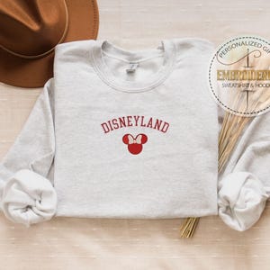 May include: A gray sweatshirt with the text "DISNEYLAND" in red embroidered on the front. A red Mickey Mouse head with a bow is embroidered below the text.