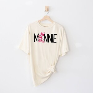 May include: A cream colored t-shirt with a black and pink Minnie Mouse graphic and the text "MINNIE" printed on the front.