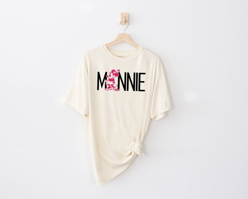 May include: A cream colored t-shirt with a black and pink Minnie Mouse graphic and the text "MINNIE" printed on the front.
