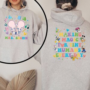 Puede incluir: Una sudadera con capucha gris claro con un dise&ntilde;o colorido. La parte delantera presenta una cabeza de Mickey Mouse con un castillo, &uacute;tiles escolares y el texto "MRS. BROWN". La parte trasera muestra personajes de Disney y el texto "MAKING MAGIC FOR TINY HUMANS EVERY DAY".
