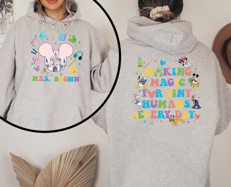 Puede incluir: Una sudadera con capucha gris claro con un dise&ntilde;o colorido. La parte delantera presenta una cabeza de Mickey Mouse con un castillo, &uacute;tiles escolares y el texto "MRS. BROWN". La parte trasera muestra personajes de Disney y el texto "MAKING MAGIC FOR TINY HUMANS EVERY DAY".