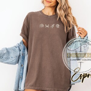May include: Brown t-shirt with embroidered sea life designs, including coral, a crab, a seashell, and a scallop. The shirt is a comfort colors style. The text "Embroidered Comfort Colors" is visible.