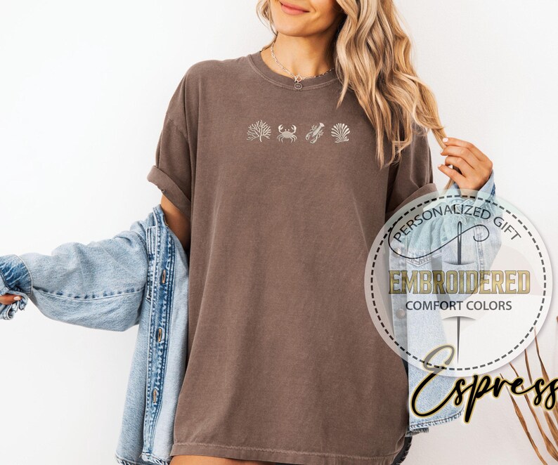 May include: Brown t-shirt with embroidered sea life designs, including coral, a crab, a seashell, and a scallop. The shirt is a comfort colors style. The text "Embroidered Comfort Colors" is visible.