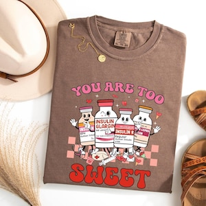 May include: A brown t-shirt with the text "YOU ARE TOO SWEET" in pink and red. The shirt features a cartoon graphic of insulin bottles with arms and legs. A gold necklace and brown sandals are also visible.