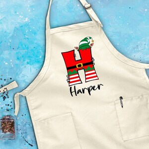 Puede incluir: Delantal de cocina blanco con una letra H roja y verde decorada con un duende de Navidad. El nombre "Harper" est&aacute; impreso debajo de la letra.