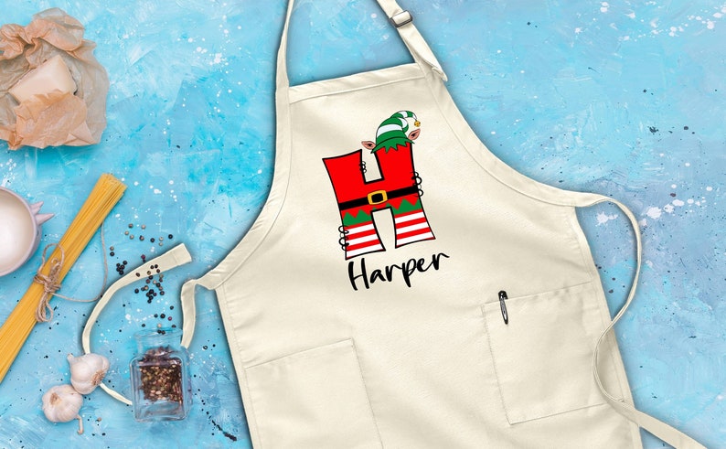 Puede incluir: Delantal de cocina blanco con una letra H roja y verde decorada con un duende de Navidad. El nombre "Harper" est&aacute; impreso debajo de la letra.