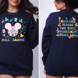 Puede incluir: Sudadera azul marino con un dise&ntilde;o colorido con tem&aacute;tica de Disney. La parte delantera presenta una cabeza de Mickey Mouse con un castillo, &uacute;tiles escolares y el nombre "Mrs. Brown". La parte trasera dice "Making Magic for Tiny Humans Every Day" con personajes de Disney.