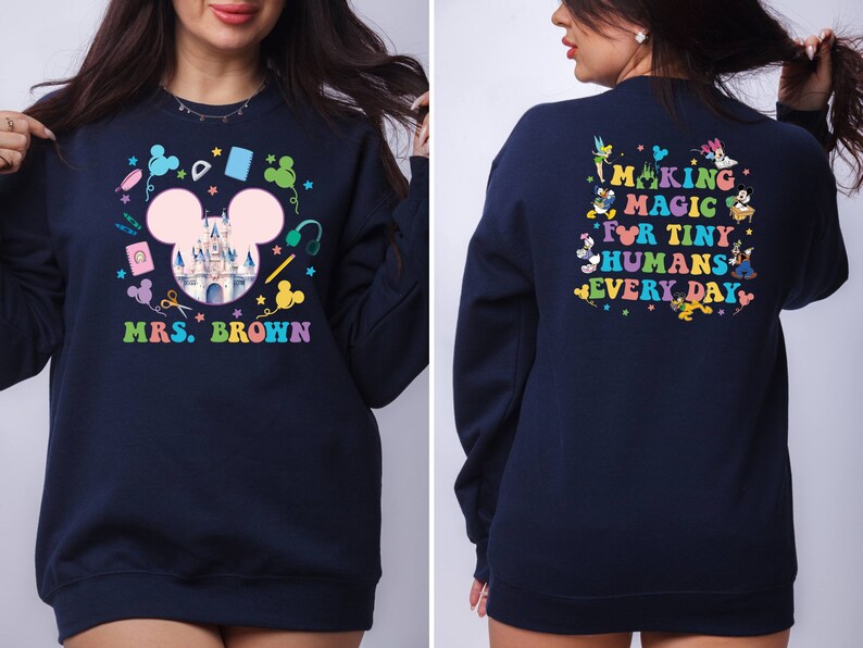 Puede incluir: Sudadera azul marino con un dise&ntilde;o colorido con tem&aacute;tica de Disney. La parte delantera presenta una cabeza de Mickey Mouse con un castillo, &uacute;tiles escolares y el nombre "Mrs. Brown". La parte trasera dice "Making Magic for Tiny Humans Every Day" con personajes de Disney.