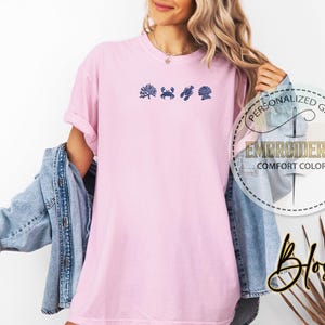 May include: A light pink t-shirt with embroidered navy blue sea-themed designs. The shirt features a coral, crab, seashell, and a scallop. The shirt has the words "PERSONALIZED GIFT" and "EMBROIDERED COMFORT COLORS" in a circular design.