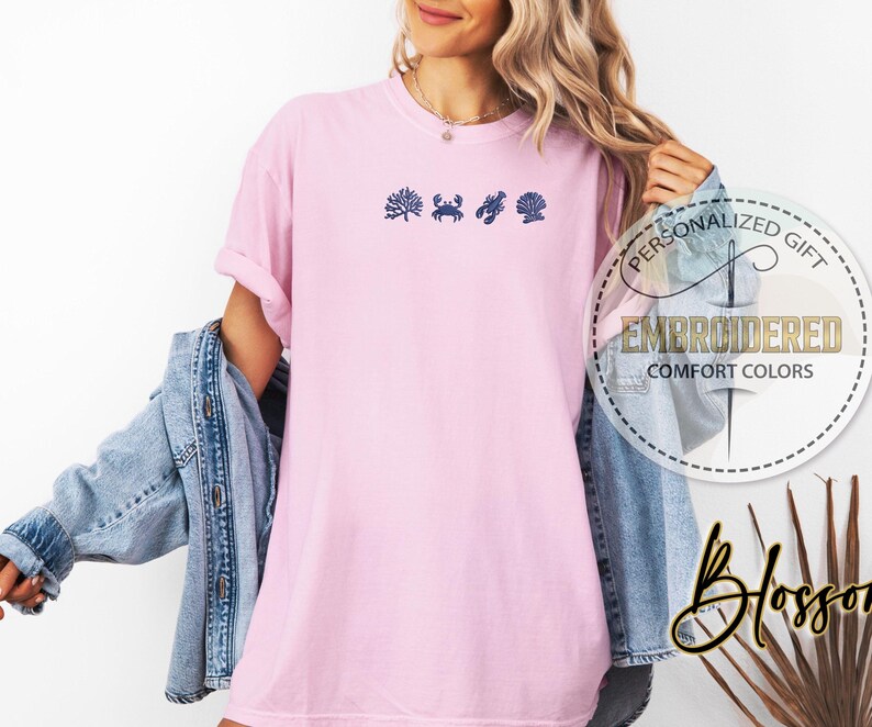 May include: A light pink t-shirt with embroidered navy blue sea-themed designs. The shirt features a coral, crab, seashell, and a scallop. The shirt has the words "PERSONALIZED GIFT" and "EMBROIDERED COMFORT COLORS" in a circular design.
