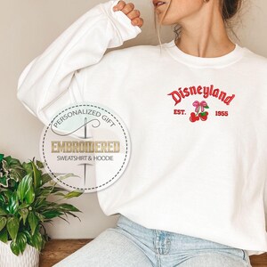 May include: White sweatshirt with a red embroidered Disneyland logo and cherries. The logo says "Disneyland" with "Est. 1955" underneath.