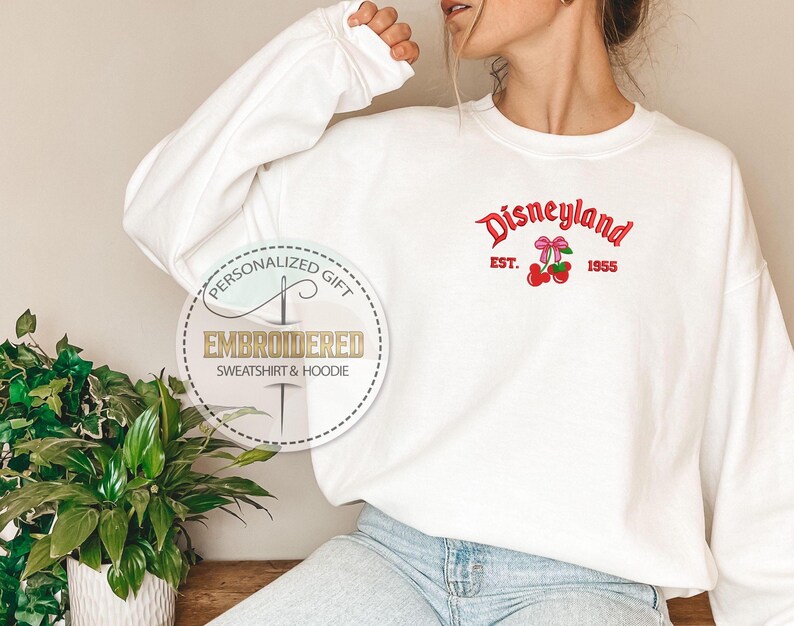 May include: White sweatshirt with a red embroidered Disneyland logo and cherries. The logo says "Disneyland" with "Est. 1955" underneath.