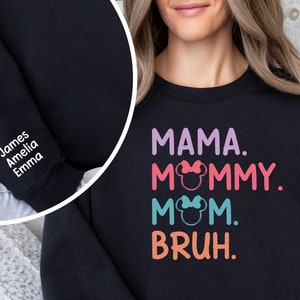 K&ouml;nnte beinhalten: Schwarzer Pullover mit dem Text "MAMA. MOMMY. MOM. BRUH." in bunten Buchstaben mit einem Mickey-Maus-Kopf-Motiv auf jedem Wort. Der Pullover hat auch einen wei&szlig;en bestickten &Auml;rmel mit den Namen "James, Amelia, Emma".