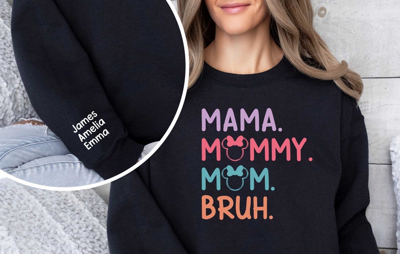 K&ouml;nnte beinhalten: Schwarzer Pullover mit dem Text "MAMA. MOMMY. MOM. BRUH." in bunten Buchstaben mit einem Mickey-Maus-Kopf-Motiv auf jedem Wort. Der Pullover hat auch einen wei&szlig;en bestickten &Auml;rmel mit den Namen "James, Amelia, Emma".