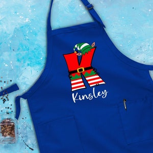 Puede incluir: Delantal azul de Navidad con un dise&ntilde;o de elfo rojo y verde y el nombre "Kinsley" impreso en blanco.