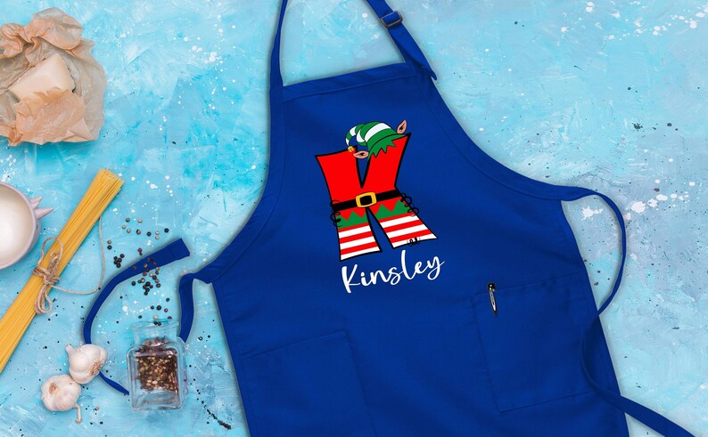 Puede incluir: Delantal azul de Navidad con un dise&ntilde;o de elfo rojo y verde y el nombre "Kinsley" impreso en blanco.