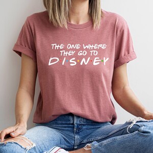 Pu&ograve; includere: T-shirt rosa polveroso con maniche arrotolate. Il testo bianco recita "The One Where They Go To Disney" con la parola "Disney" in lettere colorate. La persona indossa jeans blu.